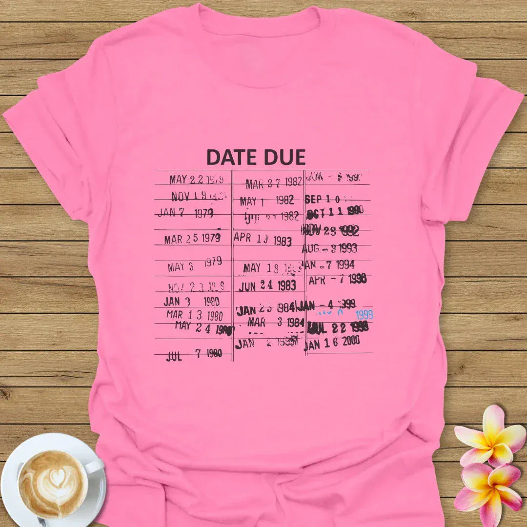 Pink t-shirt with 'DATE DUE' text and printed dates on a wooden surface with a coffee cup and flower.