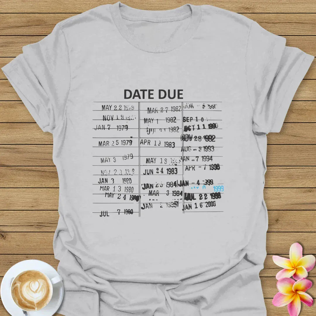 Gray t-shirt with 'DATE DUE' text on a wooden surface with a coffee cup and flower.